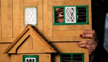 Image of a wooden model house with attractive gables, and a concealed person behind it who is peeping through a window.